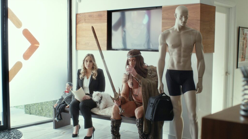 A woman in a business suit sits with a puppet and a small dog. Beside her, a man dressed as a warrior holds a sword. They sit next to a mannequin in black underwear.